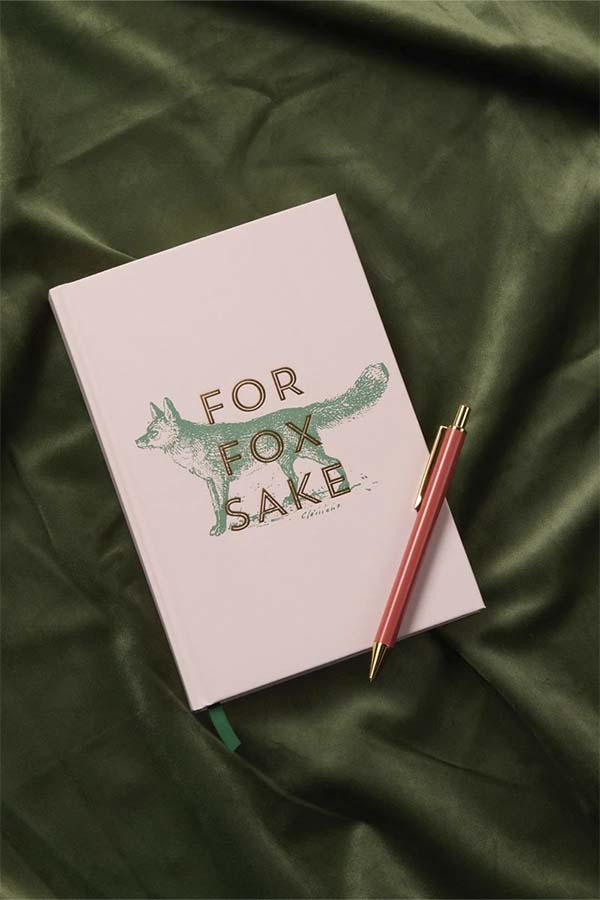 Cuaderno de tapa dura en tono beige con la frase "For Fox Sake" acompañada de una ilustración de zorro en verde y dorado. Incluye marcapáginas de cinta verde y páginas diseñadas para notas y planificación.